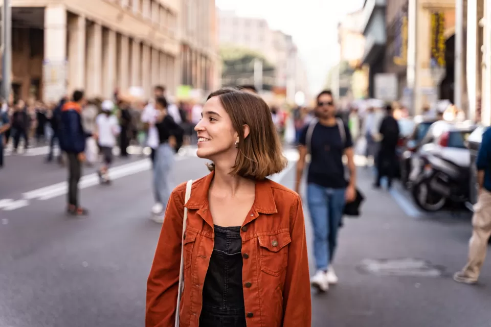 Brown-haired woman in an orange jacket walking down crowded street smiling